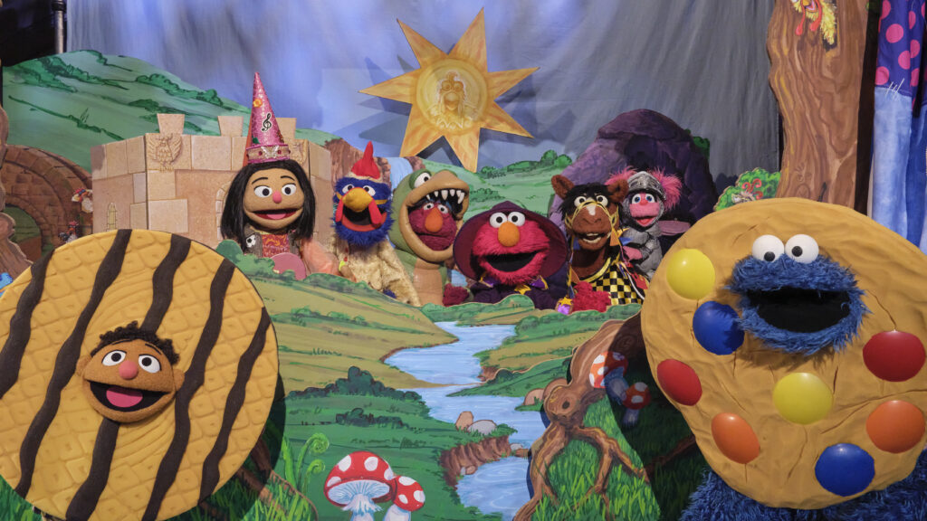 "Colorful puppets in costume pose in a whimsical fantasy landscape with a castle, bright sun, river, and giant cookies, evoking a playful, magical, and storybook setting reminiscent of a puppet show."