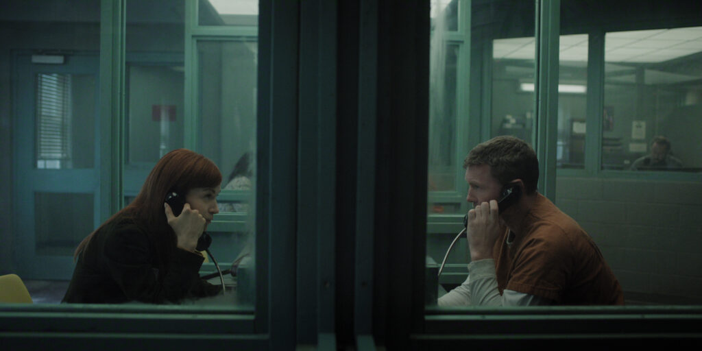 "A woman and a man in prison uniform sit across glass in a visitation room using phones to talk. The setting is dimly lit with institutional green walls and other people are visible in the background."